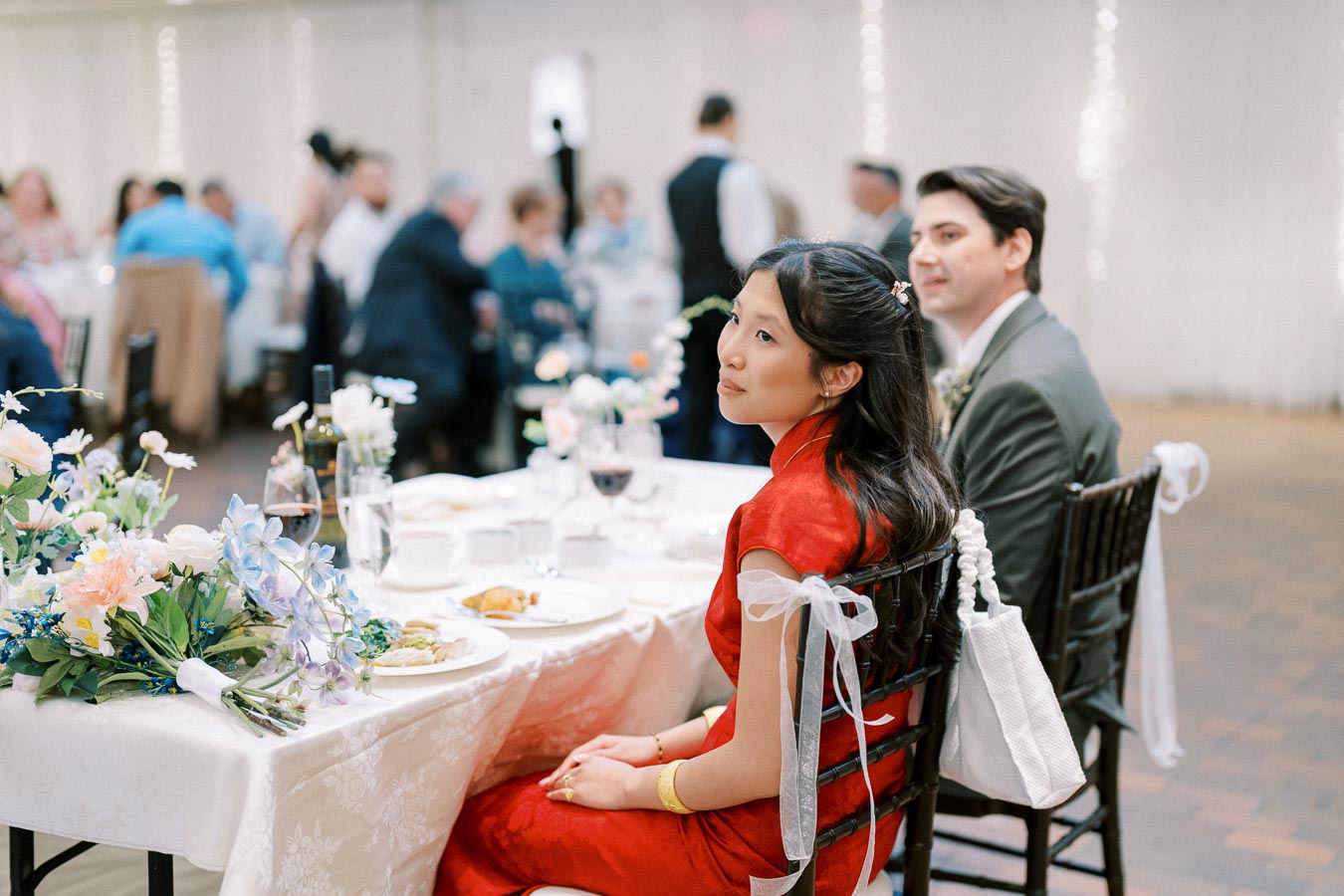 Elegant wedding reception with guests seated at a beautifully decorated table featuring floral arrangements and fine dining settings, focusing on a woman in a red dress and a man in a suit seated at the table.