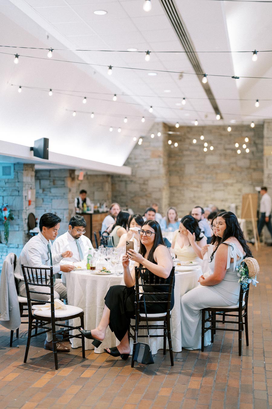 A group of elegantly dressed wedding guests seated at a round table in a beautifully decorated reception hall with string lights and stone walls.