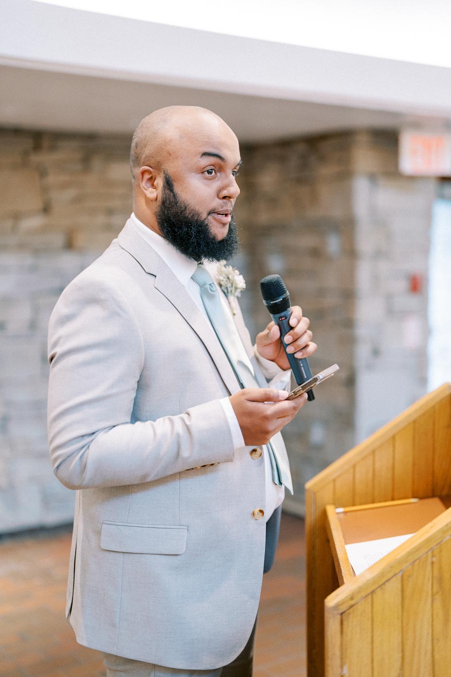 A man in a light gray suit and tie giving a speech, holding a microphone and smartphone, standing at a wooden podium with a stone wall background.