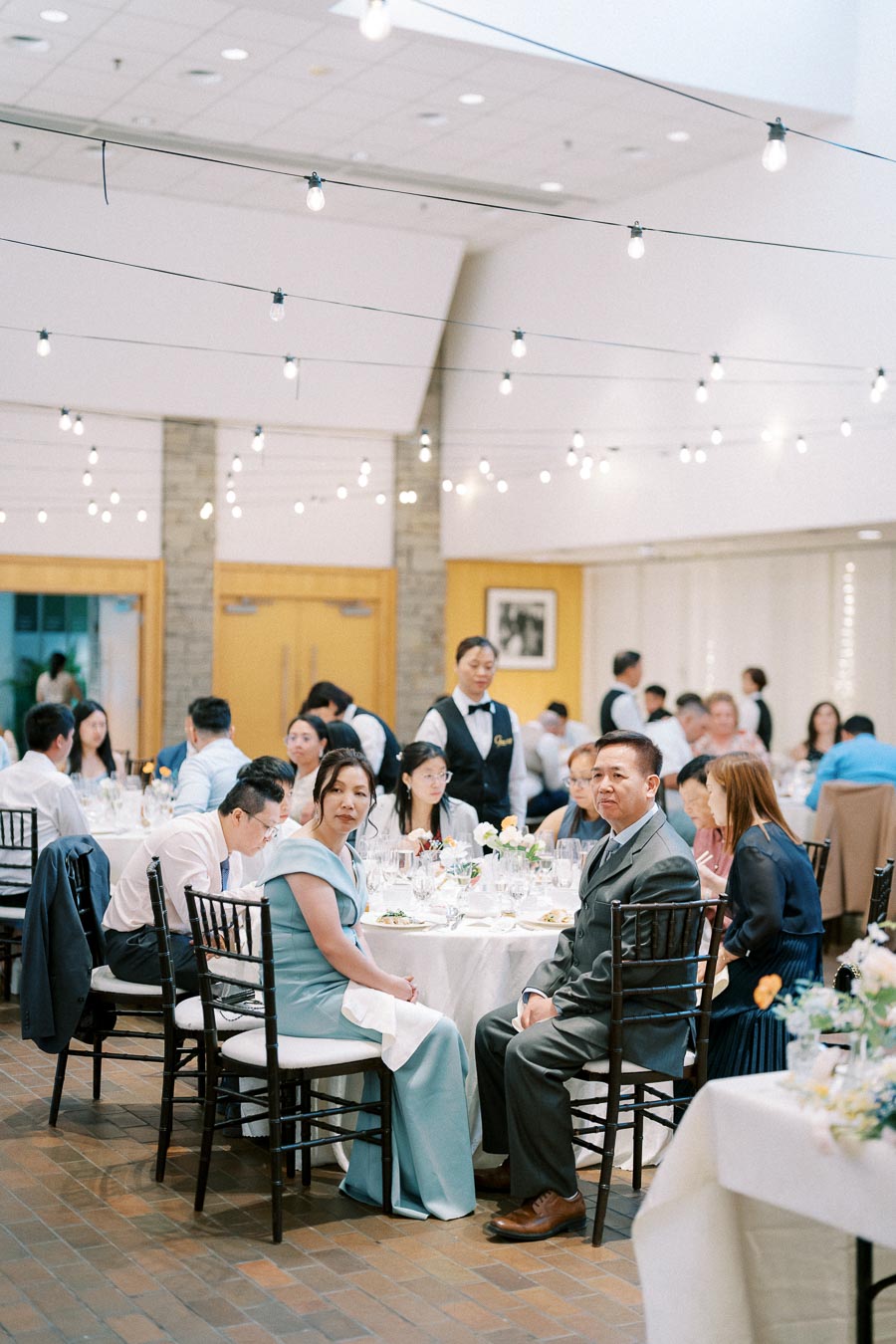 A beautifully decorated reception hall with string lights, elegantly dressed guests seated around a round table, and a waiter serving the guests during a formal event.