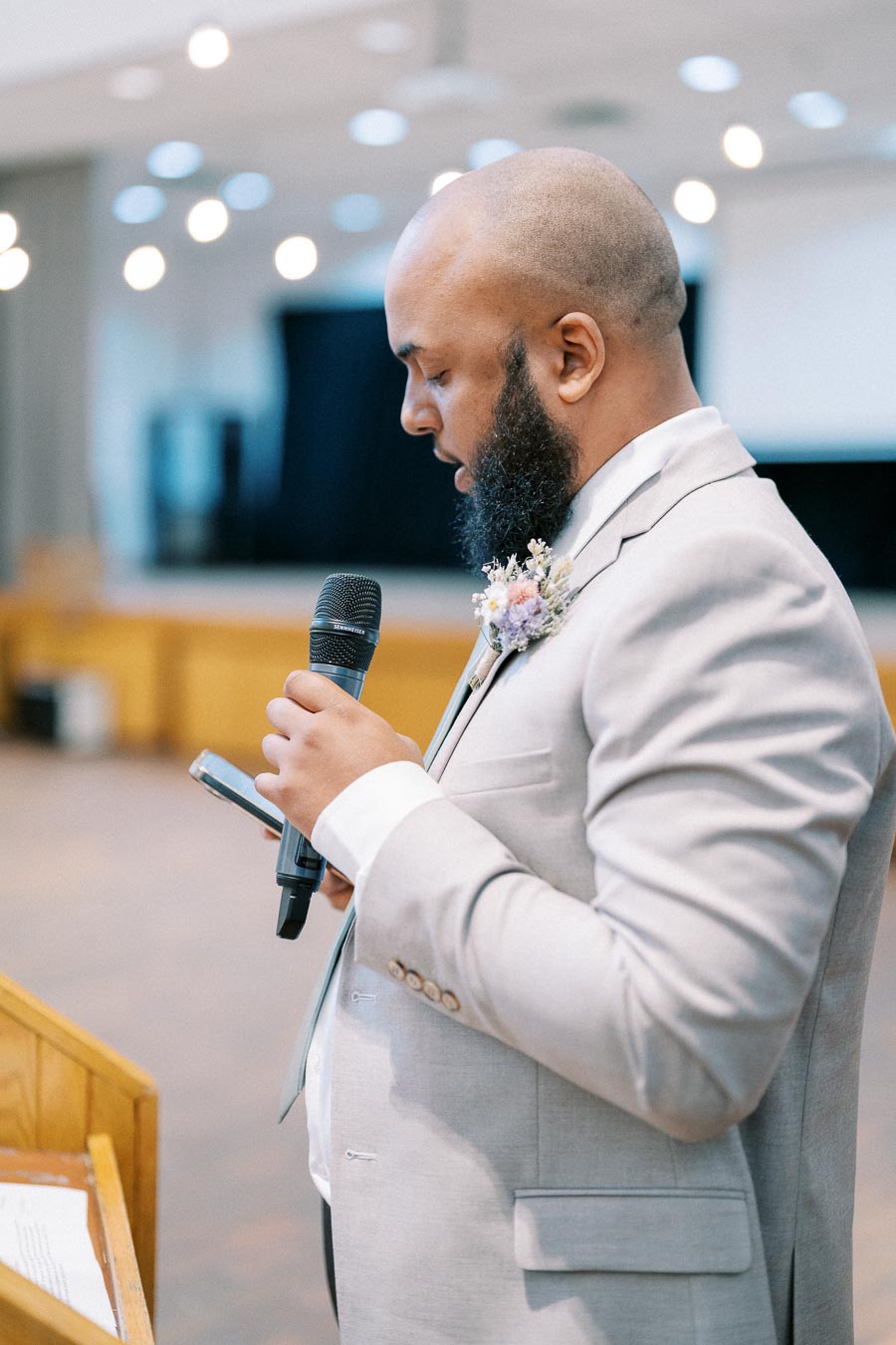 A man in a light gray suit gives a speech holding a microphone and a phone, standing at a wooden podium in a well-lit venue.