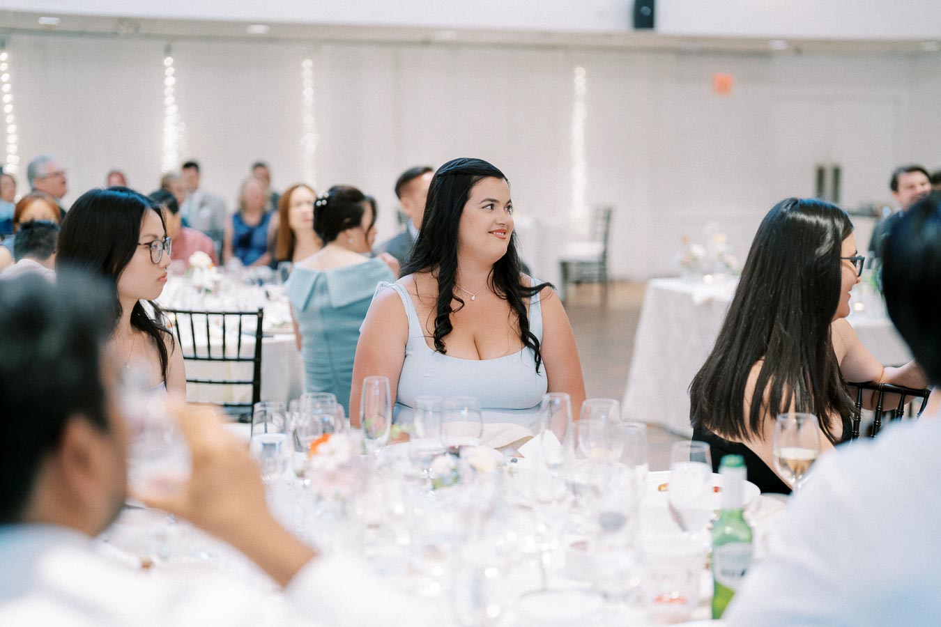 Guests seated at elegantly set tables during a formal event, with a focus on a woman in a light blue dress looking to the side, surrounded by elegantly dressed attendees, glassware, and floral centerpieces.