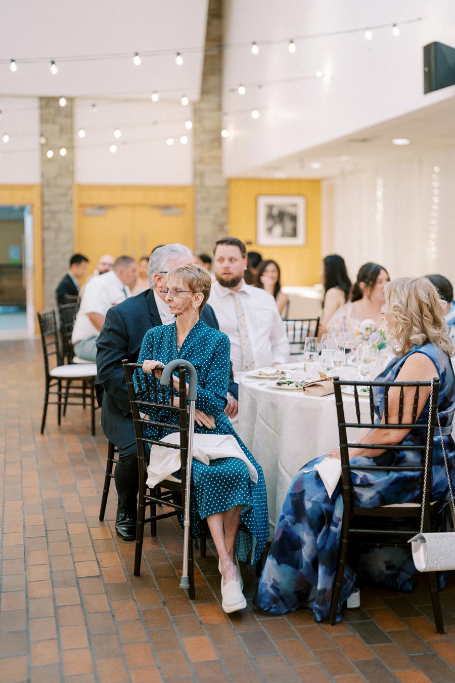Senior woman in a blue polka dot dress seated at a wedding reception table with a cane, surrounded by other guests in a warmly lit banquet hall decorated with string lights.