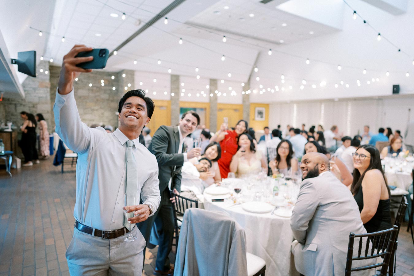 A group of people dressed in formal attire taking a selfie at a lively indoor event, with round tables and string lights creating a festive atmosphere.