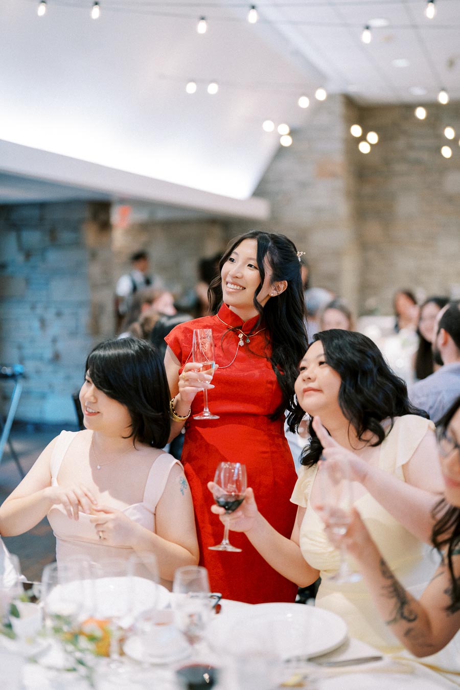 A group of three women enjoying a wedding reception, holding glasses of wine at a decorated banquet table, with string lights creating a warm ambiance.