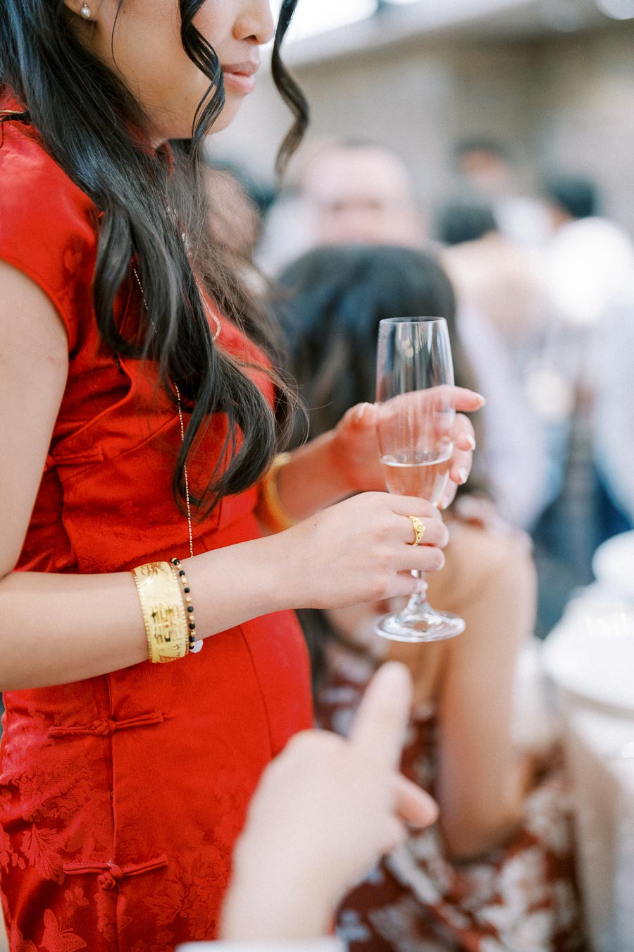 Woman in a traditional red dress holding a champagne flute at a celebration, wearing gold jewelry, with a soft-focus background of people at a gathering.