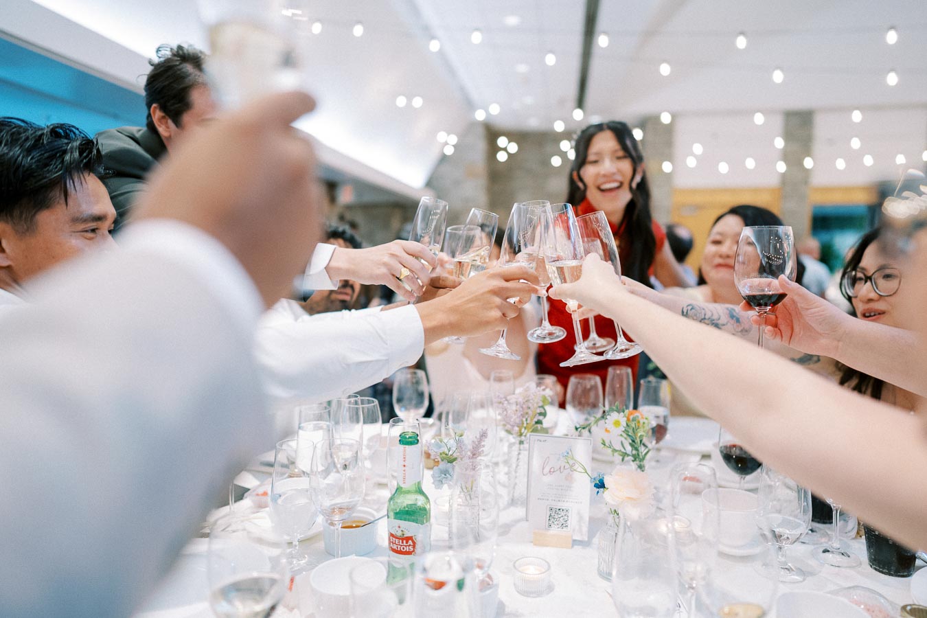 A group of people toasting with glasses at a festive celebration, seated around a decorated table with flowers and candles in a brightly lit room.