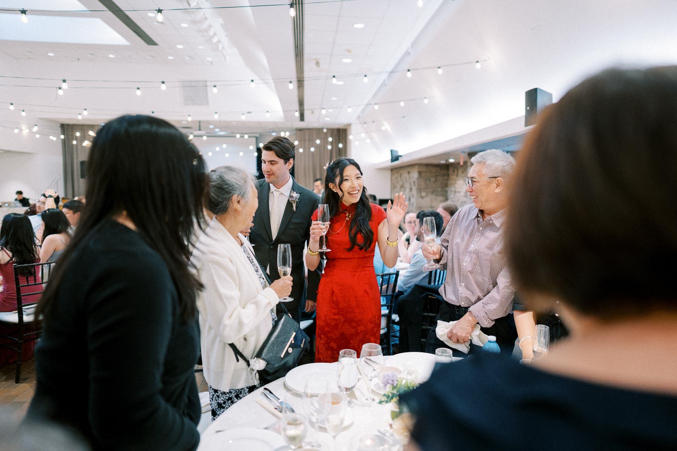 A group of people celebrating at a formal event, with a woman in a red dress smiling and holding a champagne glass, surrounded by other guests in a beautifully decorated venue with string lights and elegantly set tables.