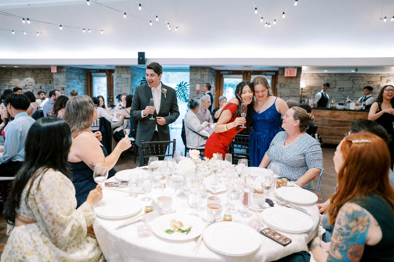 A lively wedding reception with elegantly dressed guests toasting around a decorated round table, under string lights in a cozy venue.