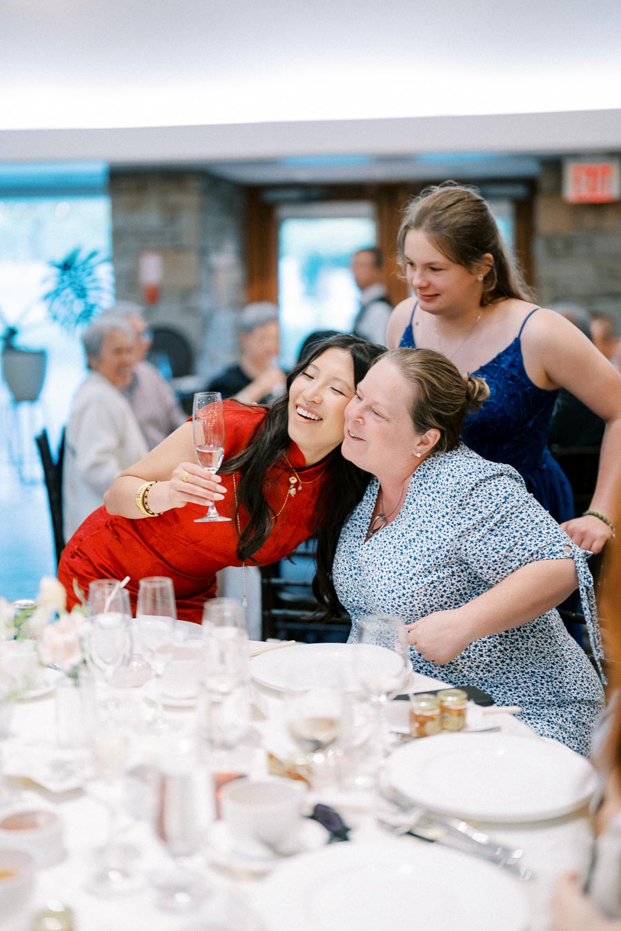 Two women smiling and celebrating at a dinner table with a drink, surrounded by elegantly set plates and glasses, in a lively banquet setting.