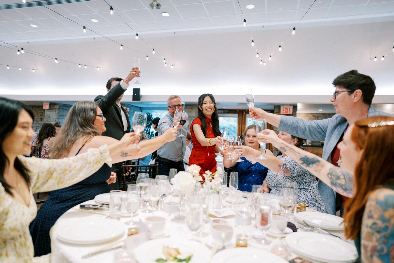 Group of people raising glasses in a celebratory toast at a lively indoor event, surrounded by elegantly set tables and ambient string lighting.