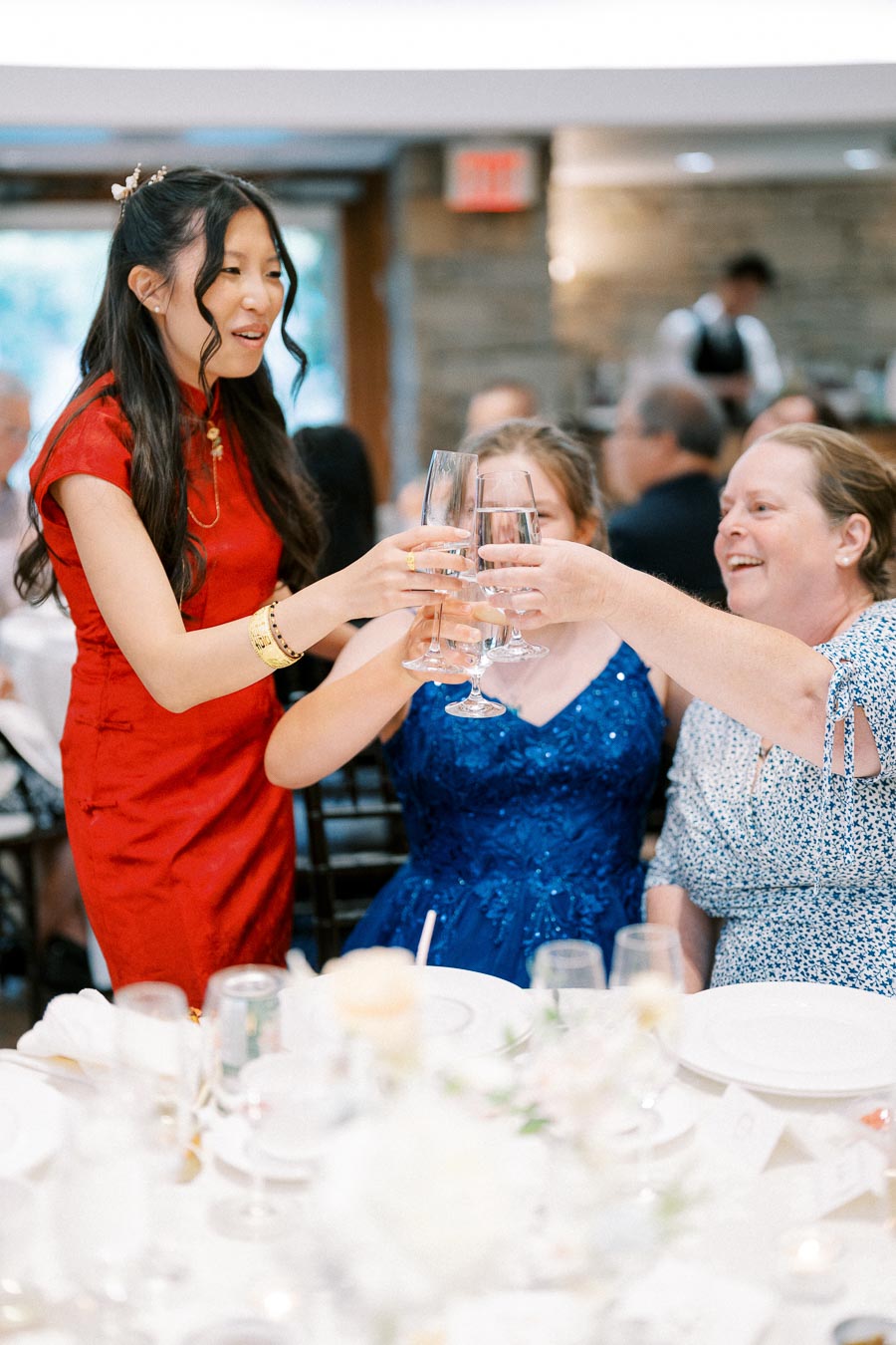 Three women celebrating and clinking glasses at a festive gathering, with one woman in a red dress and two women seated, one in a blue dress, sharing joyful moments at a decorated table.