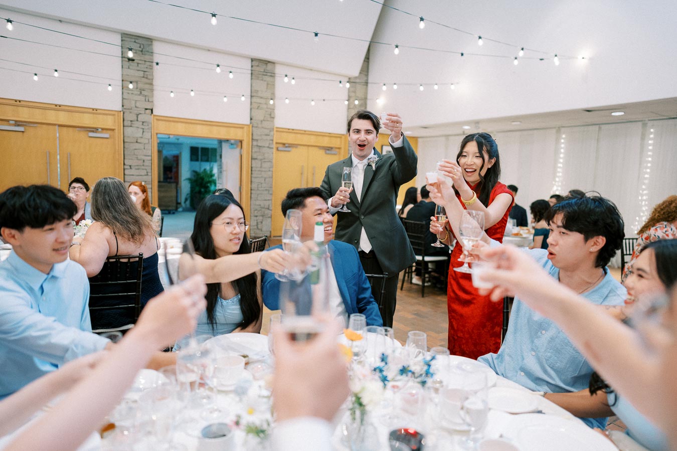 A joyful group of people raising their glasses in a celebratory toast at an indoor event, with festive string lights overhead and a warmly decorated table in the foreground.