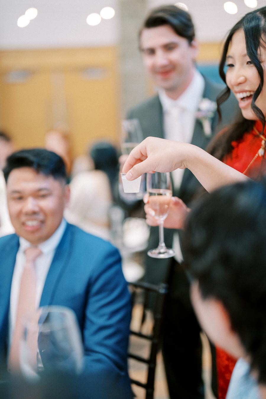 A group of well-dressed individuals are enjoying a celebration, with one person holding a glass of champagne and a small piece of paper, suggesting a toast or announcement. The room has a festive atmosphere, indicating a possible wedding or formal event.