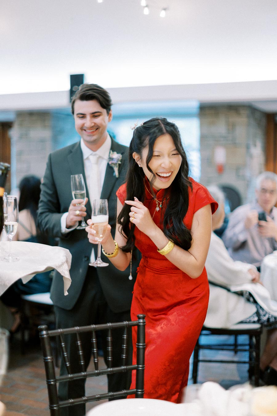 A joyful couple celebrating at a wedding reception, with the woman wearing a traditional red dress and holding a champagne glass, surrounded by guests.
