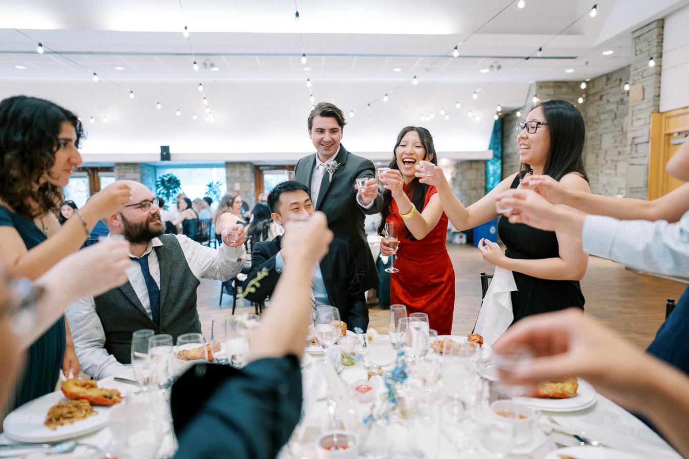 Group of people joyfully raising glasses in a toast at an indoor event with festive lighting and elegantly set tables.