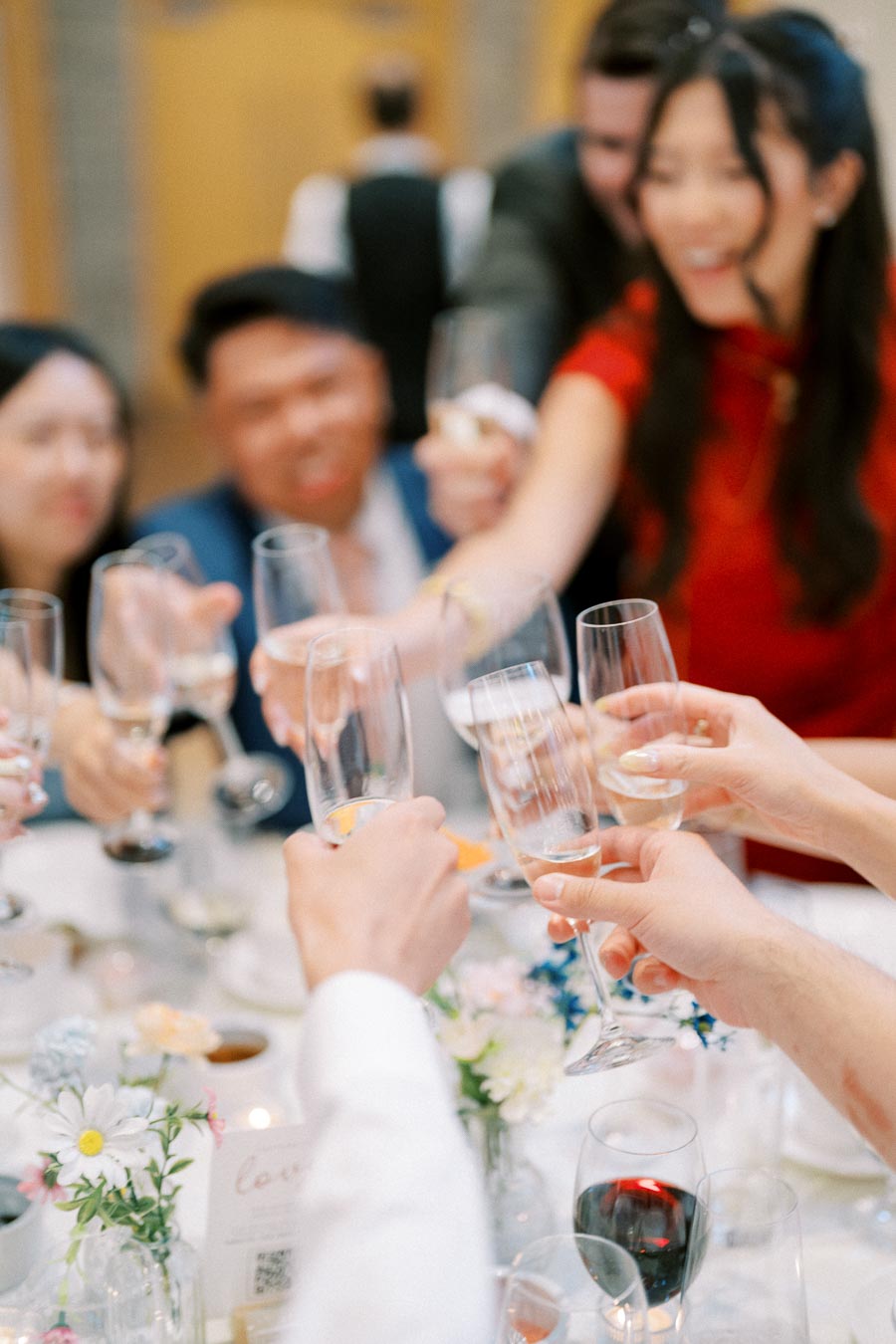 Group of people at a celebration raising champagne glasses for a toast, with blurred smiles and a festive atmosphere, flowers and wine on the table.
