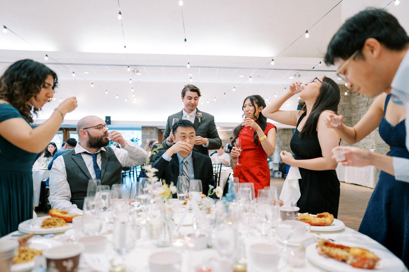 Group of people enjoying a wedding celebration, standing around a banquet table with food and drinks, dressed in formal attire.