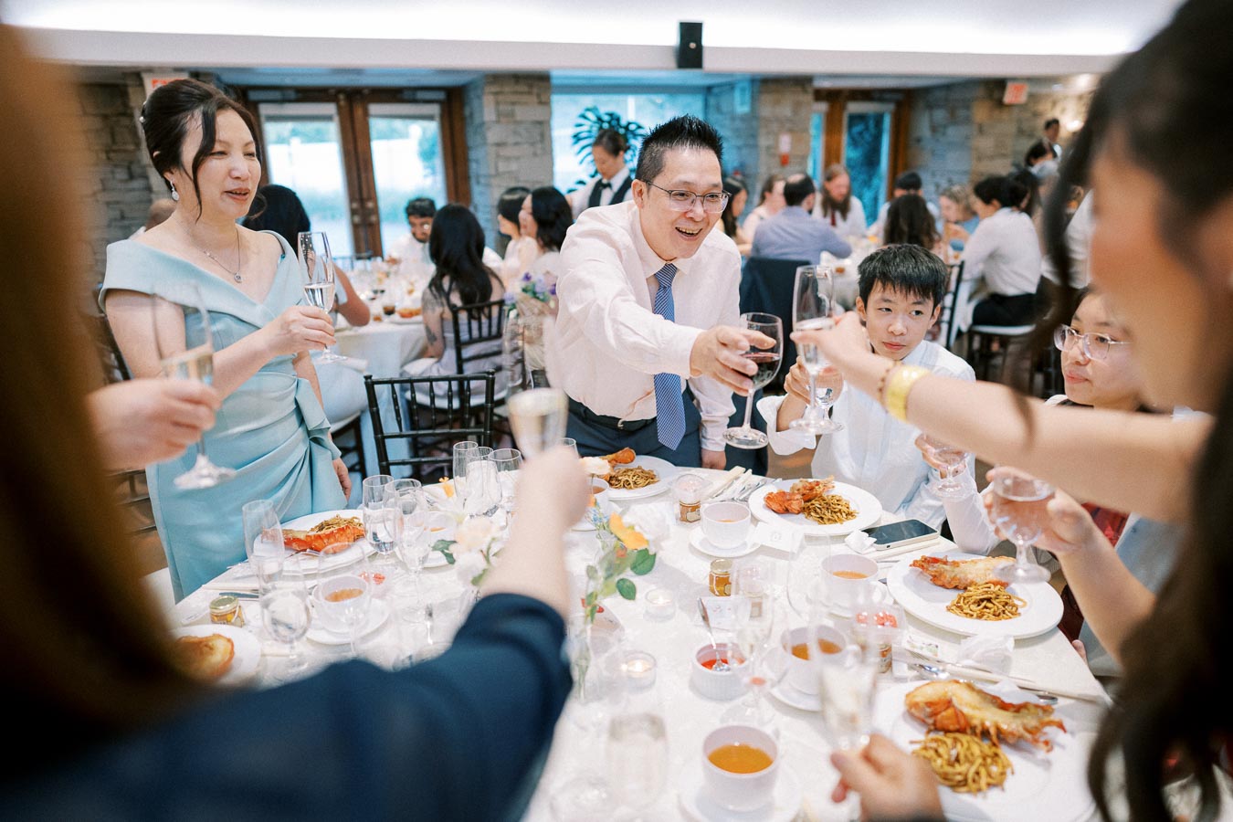 Wedding reception guests raising glasses for a toast around a table set with elaborate dishes and drinks, celebrating in a warmly lit venue.