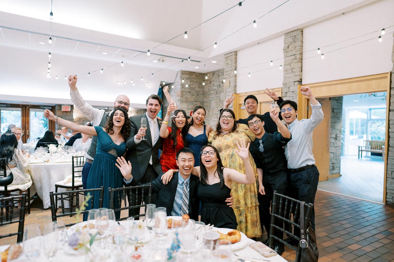 A group of people joyfully celebrating at a wedding reception, smiling and raising their arms, surrounded by elegantly set tables and festive string lights in a banquet hall.