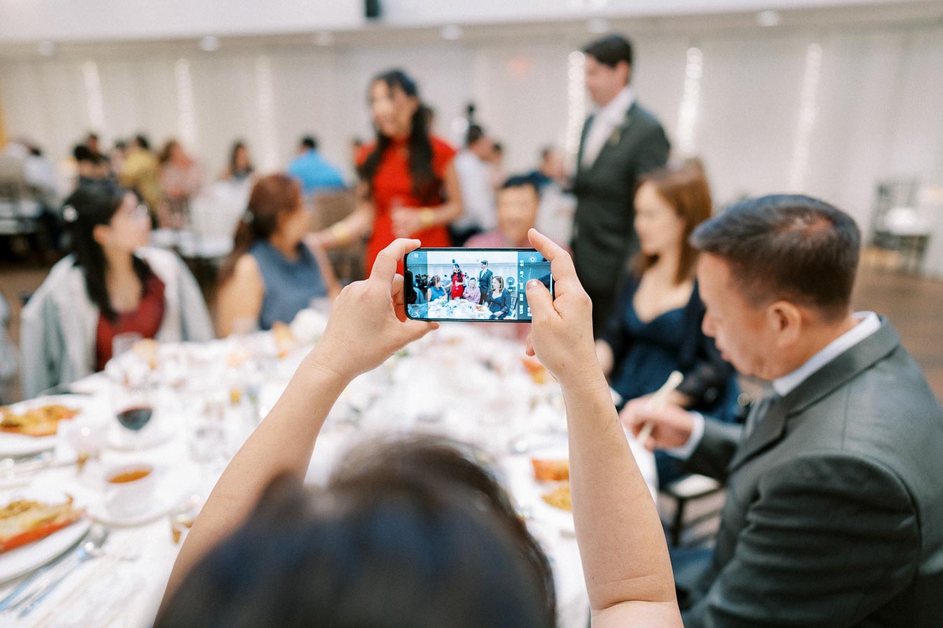 A person taking a photo with a smartphone at a family gathering or celebration, capturing a group of people sitting around a dining table, with dishes and glasses in the foreground.