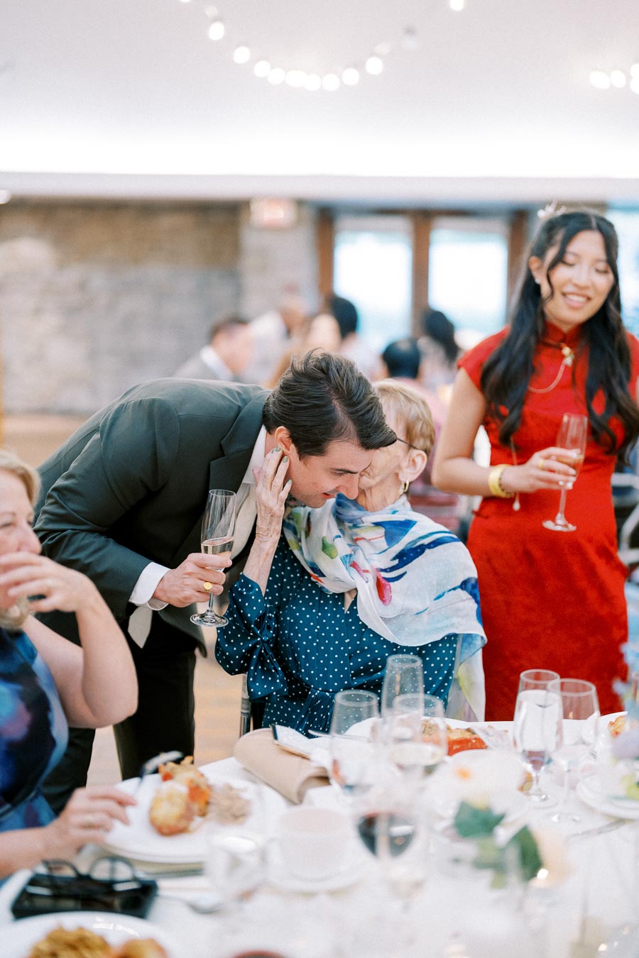 A joyful gathering at a wedding reception, featuring a man in a suit hugging an older woman with a colorful scarf at a table filled with food and drinks, while a woman in a red dress smiles holding a glass of champagne.