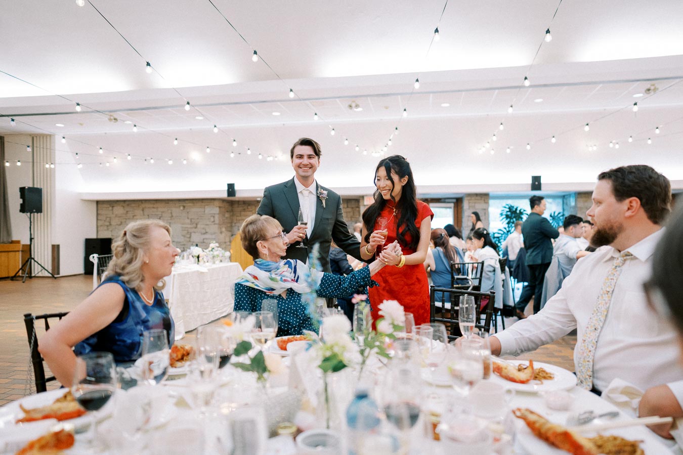 Guests enjoy a lively wedding reception with string lights overhead, while people at a table engage in conversation and celebrate with drinks.