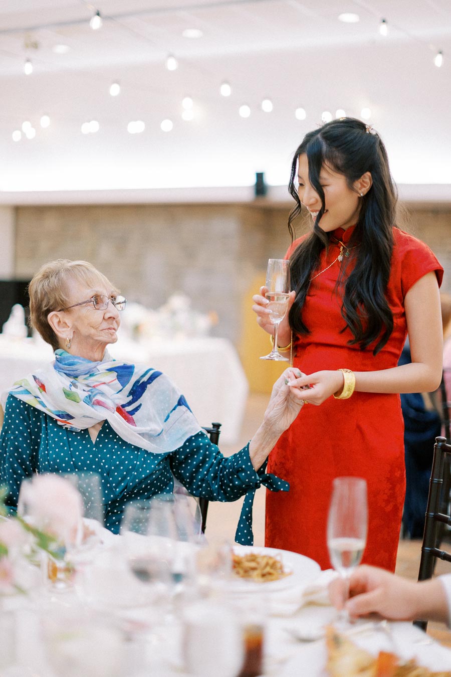 Elderly woman in teal polka dot dress and colorful scarf holding hands with a young woman in a red dress at a joyous celebration, surrounded by a softly lit, elegant dining setting.