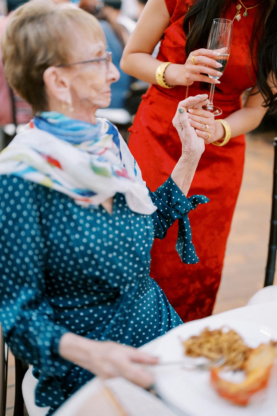 Elderly woman in a polka dot dress holding hands with a younger woman in a red dress at a social event, with a plate of food and champagne glass visible.