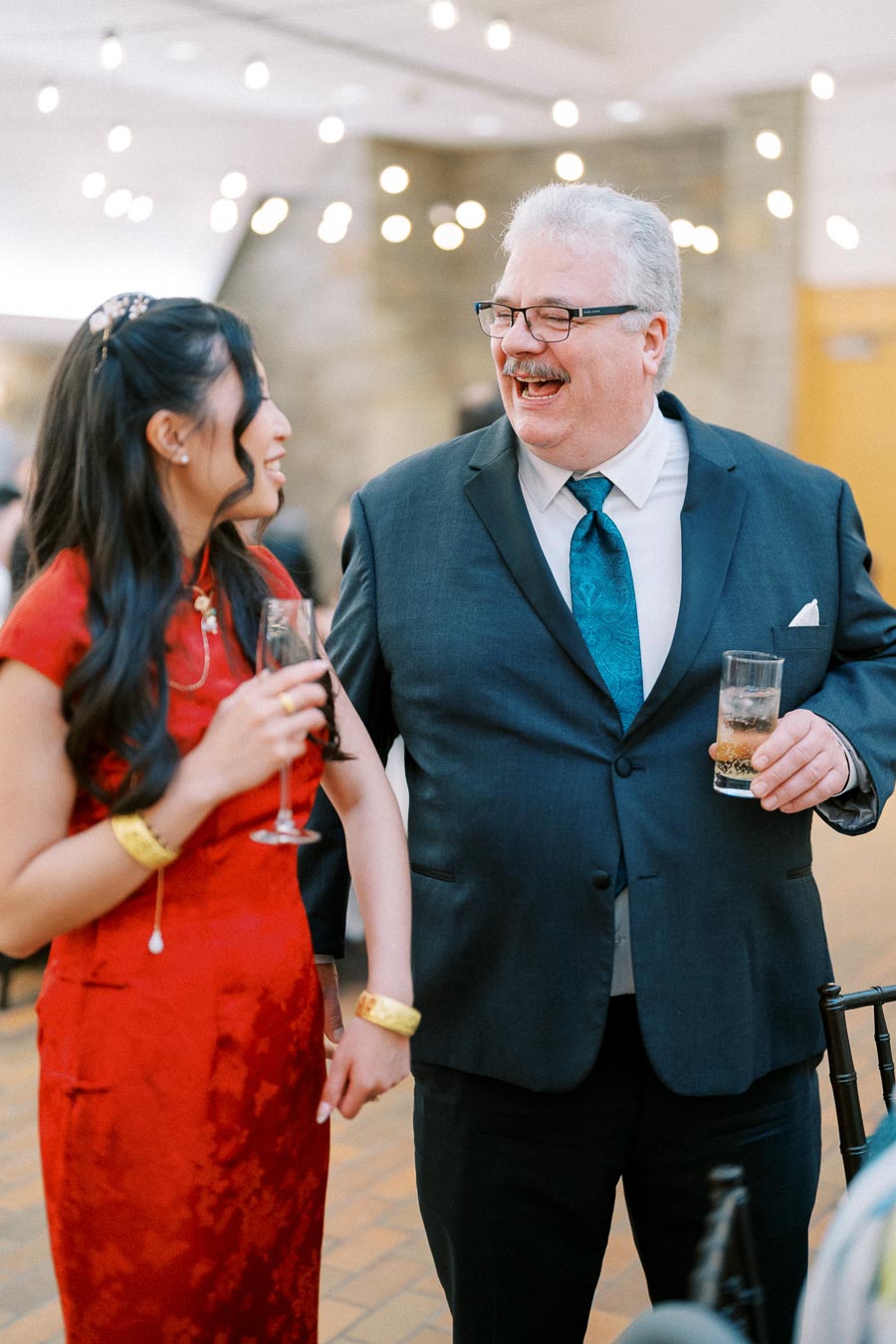 A young woman in a red dress holds hands with an older man in a suit at a festive gathering, both smiling and holding drinks under warm string lights.