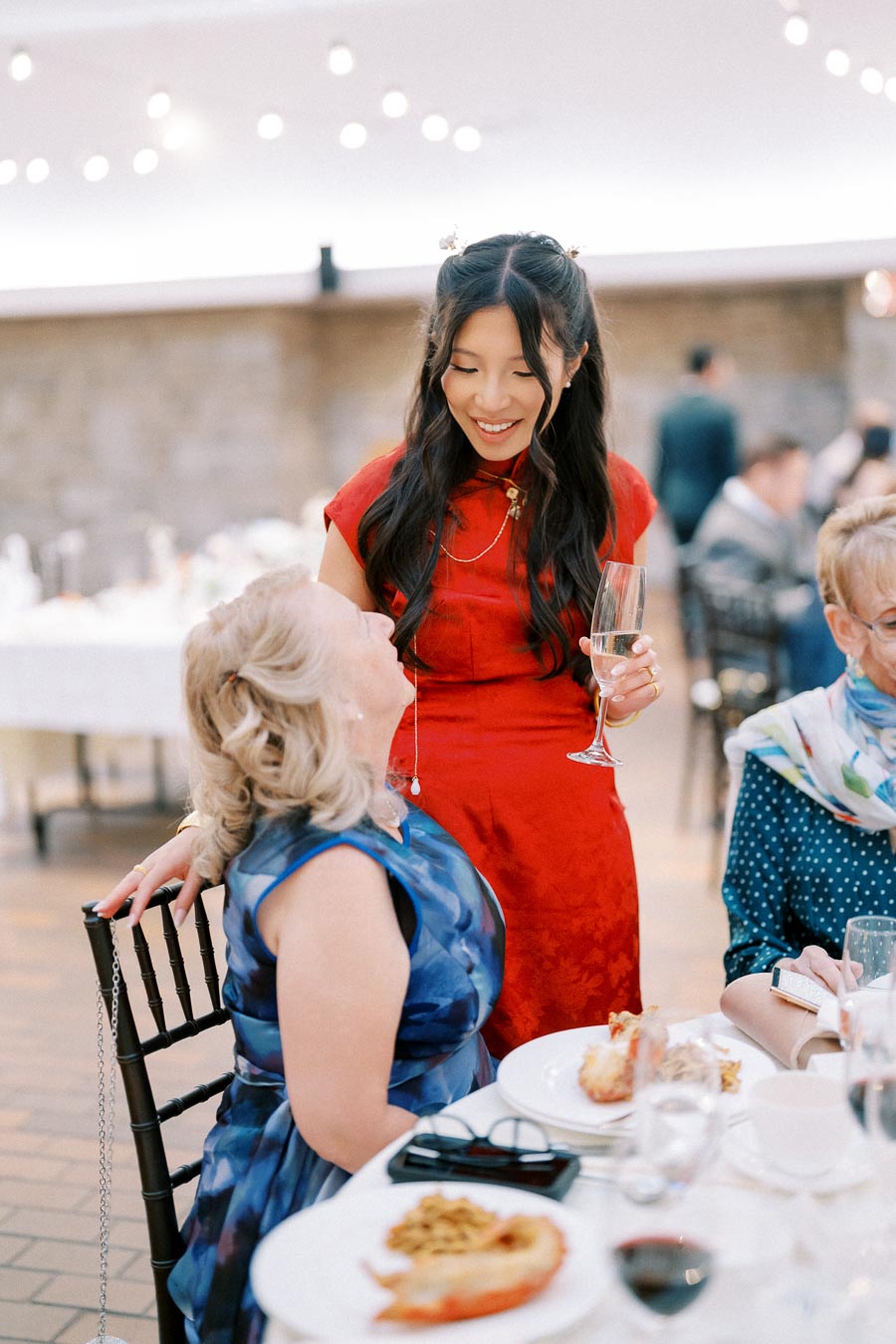 Two women smiling and chatting at a formal event, one standing in a red dress holding a champagne glass, while the other sits at a dining table with a blue dress, surrounded by elegant tableware and decorations.
