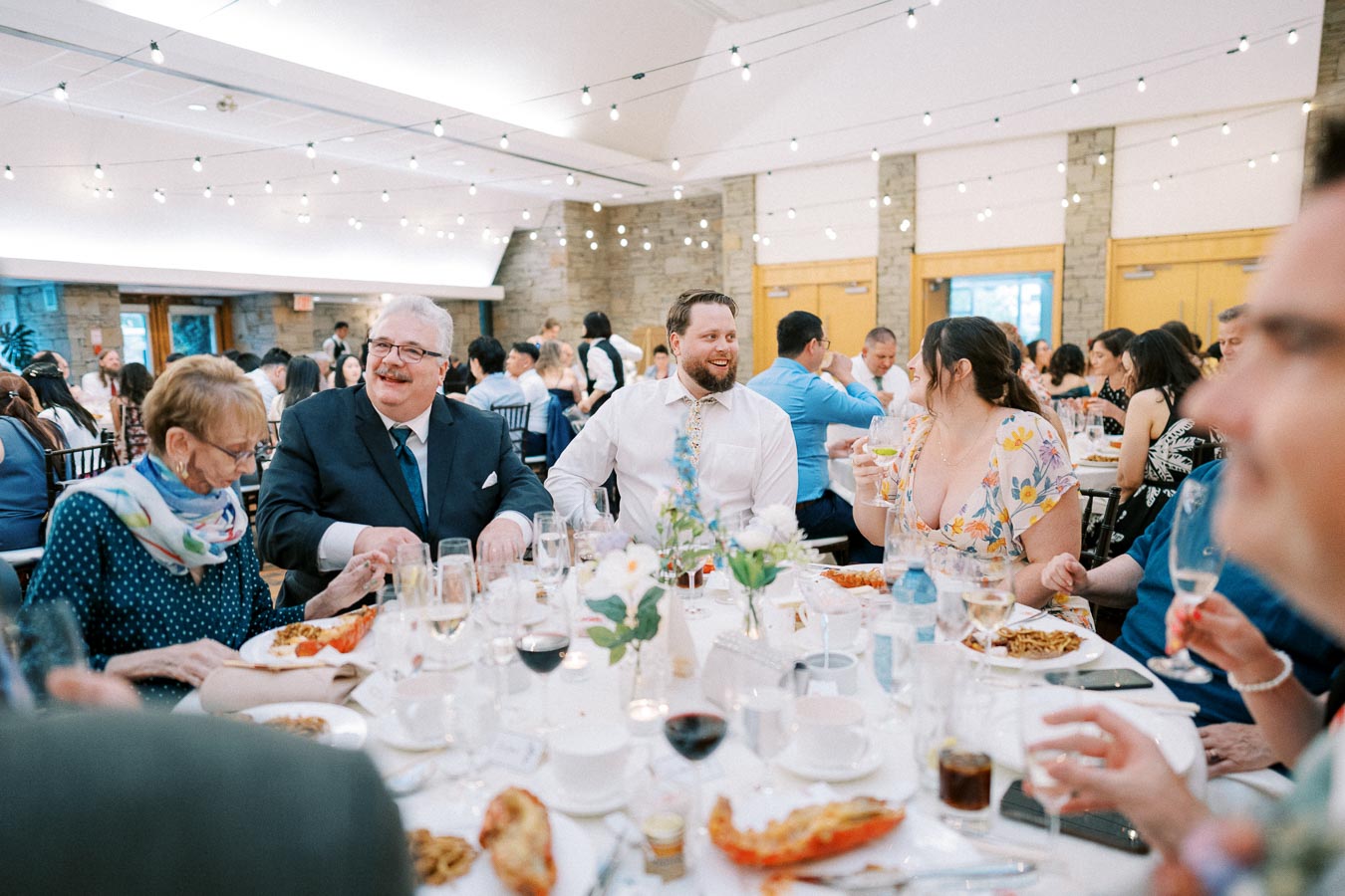 Guests enjoying a festive wedding reception at a beautifully decorated venue, featuring elegantly set tables, delicious plated meals, and twinkling string lights, creating a warm and joyful atmosphere.