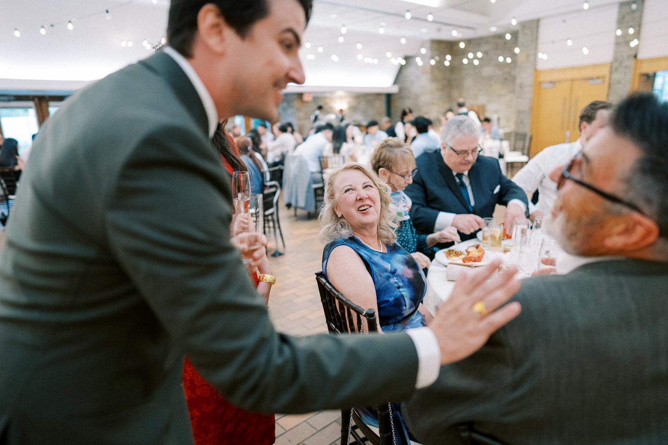 A lively wedding reception with a group of elegantly dressed people enjoying a conversation at a dinner table, ambient string lights enhancing the celebratory atmosphere.