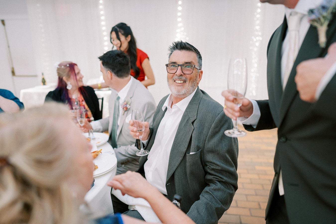A joyful man in a gray suit holds a champagne glass, surrounded by people seated at a festive gathering, with soft white lights and a relaxed atmosphere in the background.