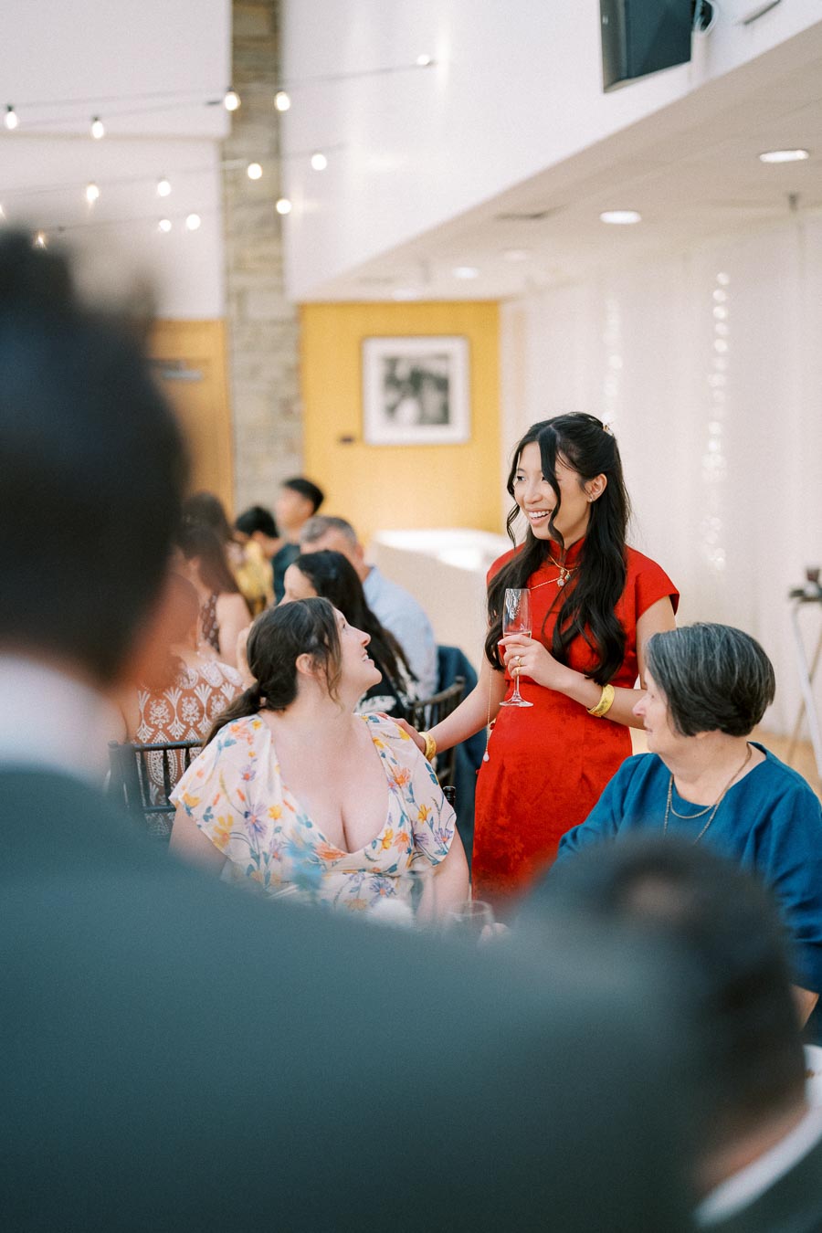 A cheerful woman in a red dress holding a champagne flute, engaging in conversation with seated guests at an elegant indoor celebration, with string lights and decor creating a festive atmosphere.
