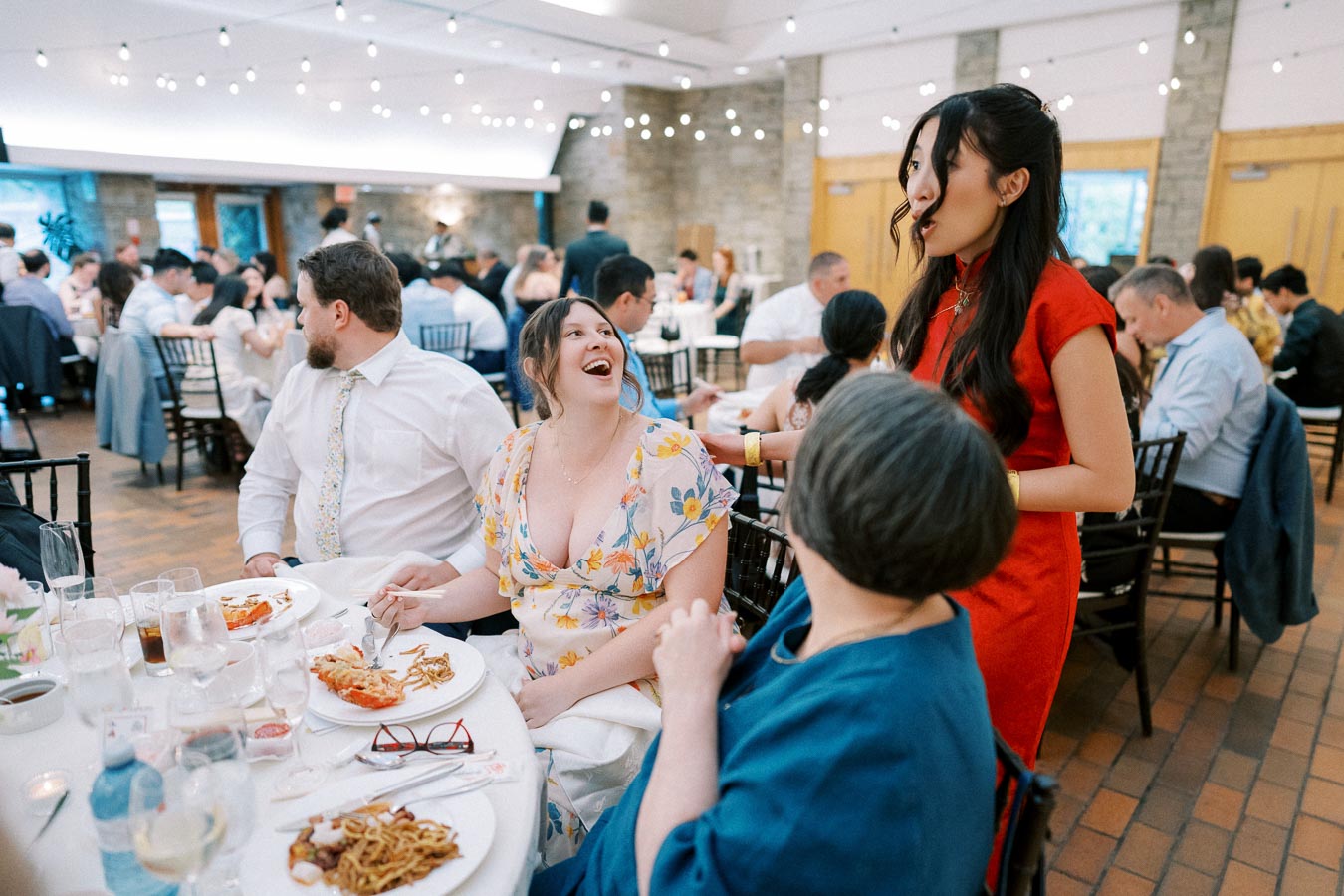 A lively wedding reception with guests seated at a decorated table, enjoying food and engaging in conversation. The room is warmly lit by string lights, creating a festive atmosphere.