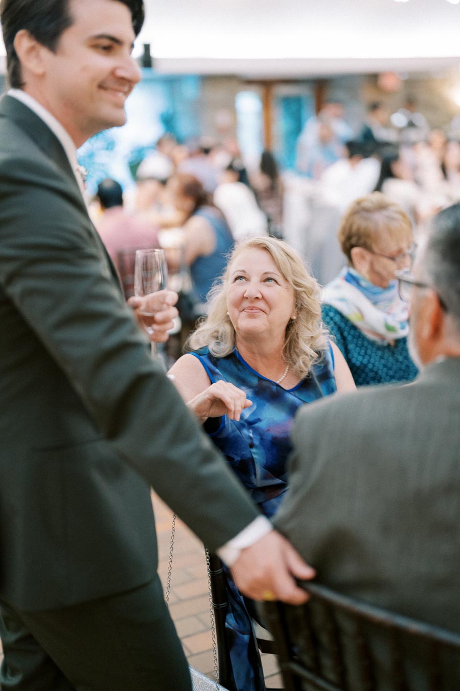 A well-dressed man holding a glass interacts with a seated woman in a blue dress during a social event, with a bustling crowd in the background.
