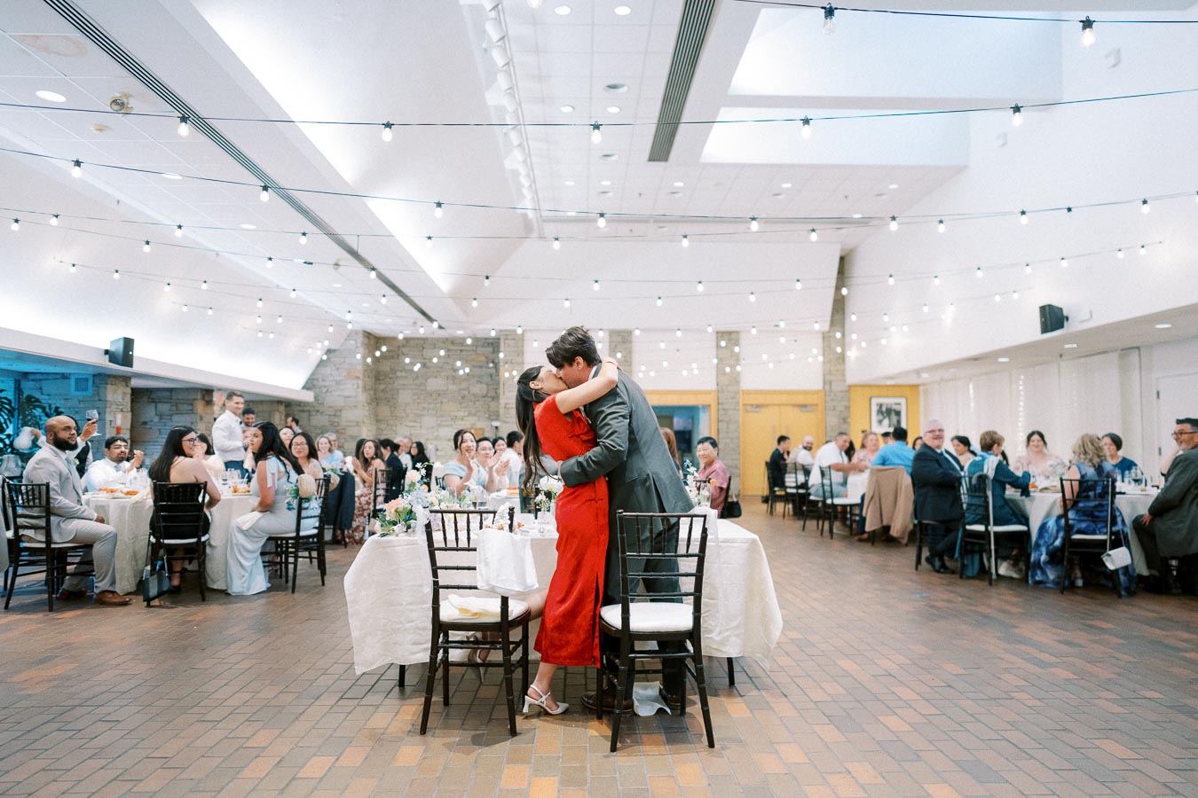 A couple embraces in the center of a warmly lit wedding reception hall with guests seated around elegantly decorated tables, under strings of twinkling lights.