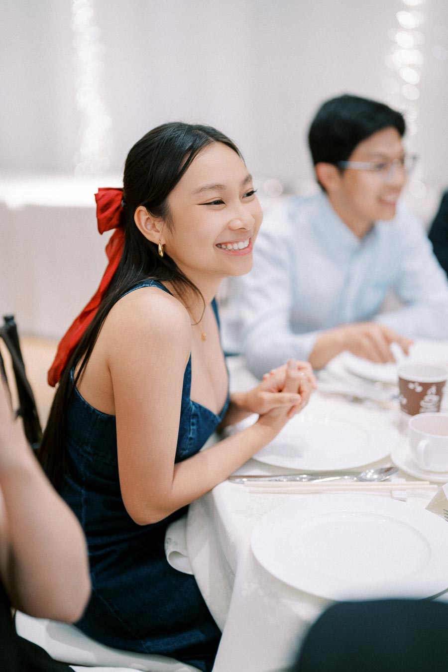 A woman with a red bow in her hair smiles while sitting at a dining table, surrounded by plates and cutlery.