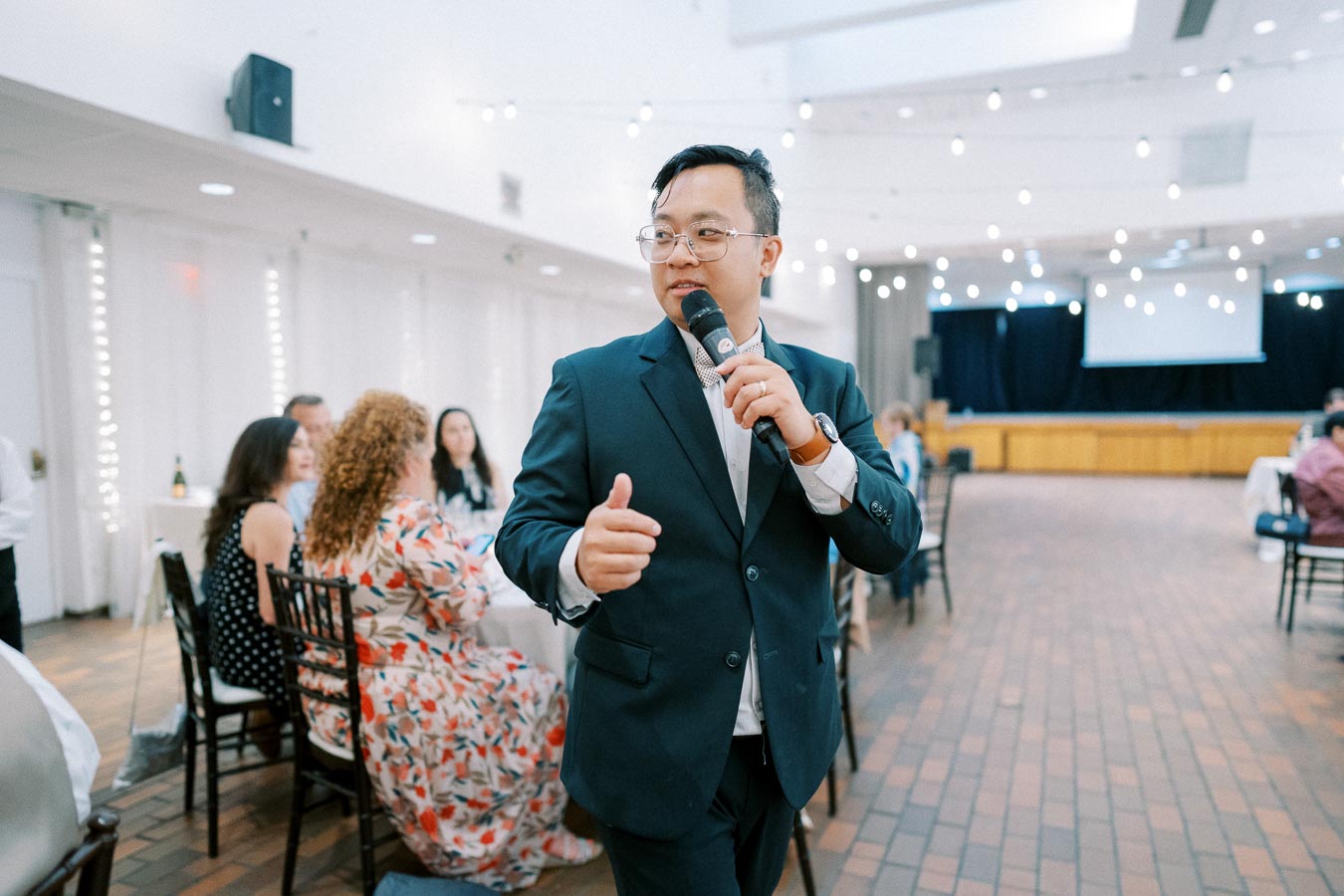 A man in a suit speaks into a microphone at a formal event, with guests seated at tables in the background.