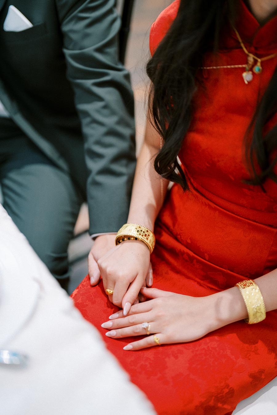 A couple holds hands, with the woman in a red dress adorned with gold jewelry, symbolizing a special occasion.
