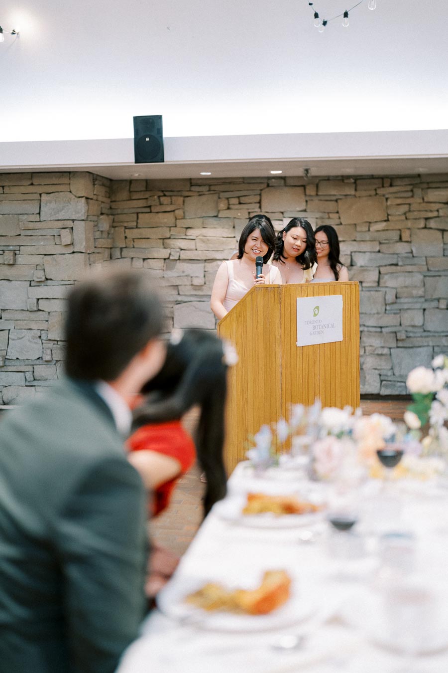 Wedding speeches being delivered by bridesmaids at a reception, with a blurred bride and groom seated at a table in the foreground, at Toronto Botanical Garden.