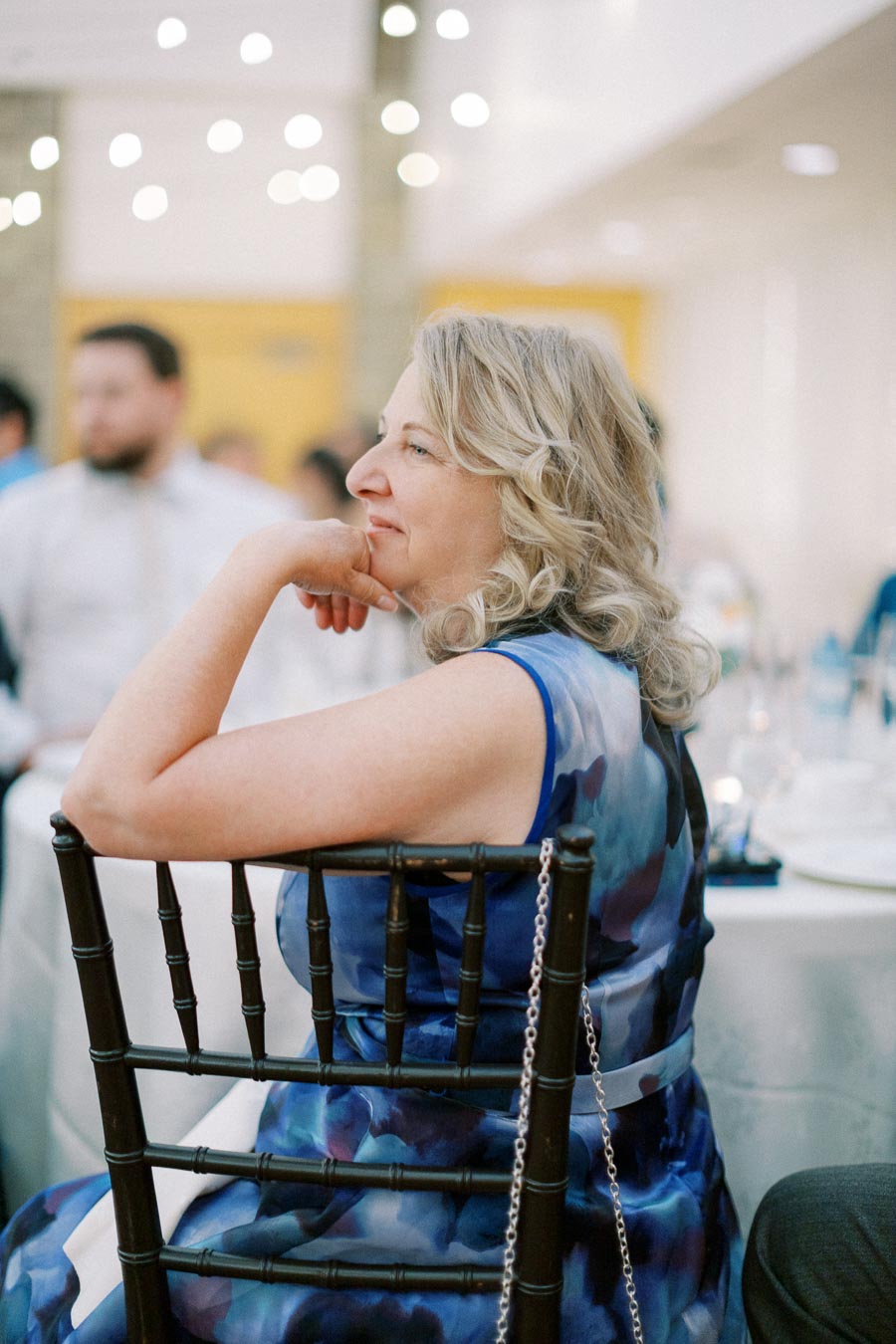 A woman in a blue dress sits attentively at an event, surrounded by softly lit decor.