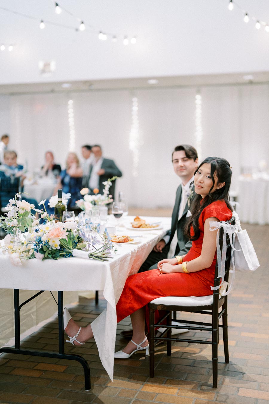 A couple seated at a wedding reception, with the woman in a red dress and the man in a suit, surrounded by floral decorations and string lights.