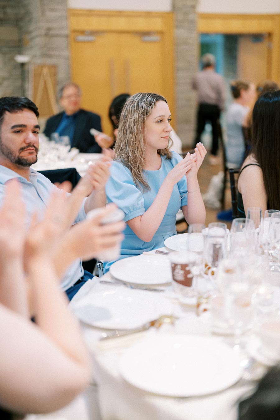 Guests at a formal event clapping while seated at a table set with plates and glasses.