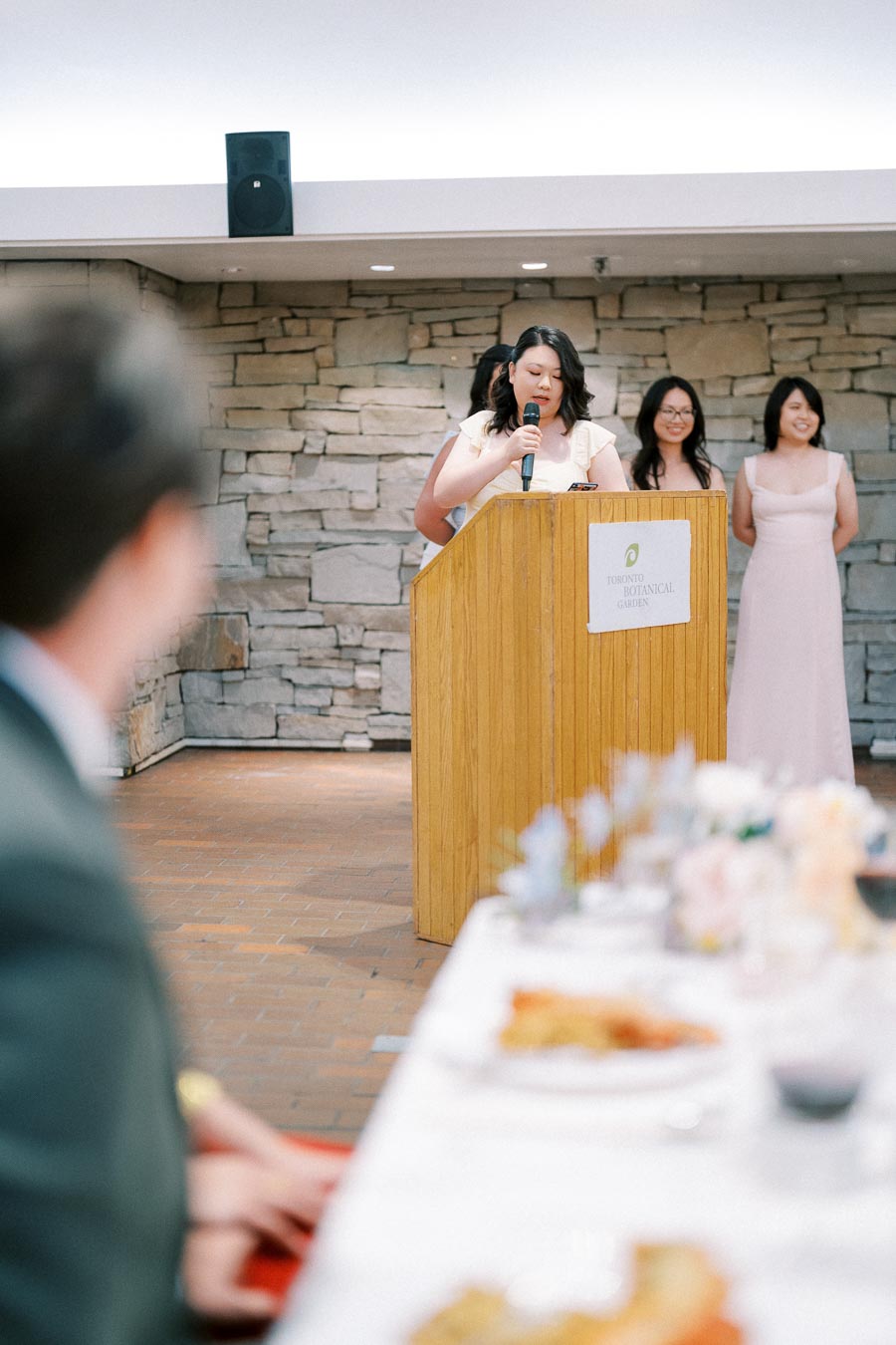 A speaker addresses guests at an event held at the Toronto Botanical Garden, with two women standing behind her. The setting features a stone wall and a table with food and drinks in the foreground.