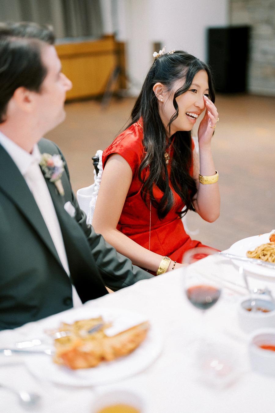A joyful woman in a red dress laughing at a formal dining event, seated beside a man in a suit, with plates of food and wine glasses on the table.