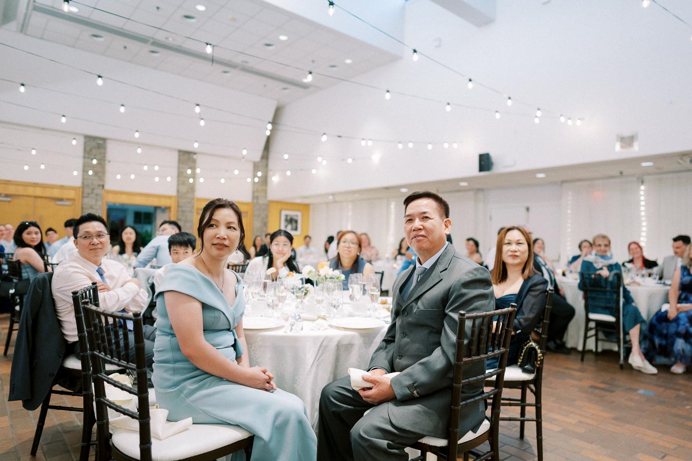 A group of well-dressed individuals seated at a banquet table during a formal event in a bright, elegantly decorated venue with string lights and high ceilings.