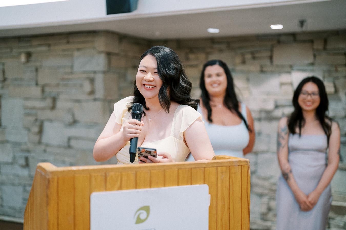 A woman delivers a speech at a podium during an event, with two other women smiling in the background.