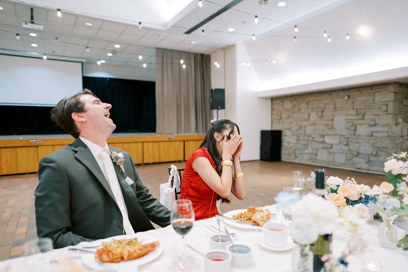 A couple sharing a joyful moment at a wedding reception. The man is laughing in a suit, while the woman in a red dress hides her face with her hands, both sitting at a table with plates of food and drinks. Decorated with flowers and soft lighting, the venue exudes a festive atmosphere.